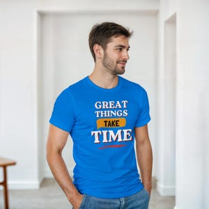 Puede incluir: Una camiseta azul con el texto "GREAT THINGS TAKE TIME" en blanco, amarillo y rojo. La camiseta es de manga corta y tiene cuello redondo.