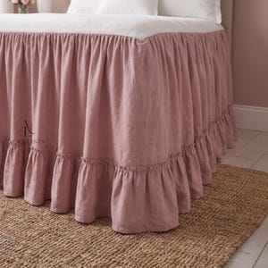 May include: A dusty rose bed skirt featuring a gathered ruffle design along the bottom edge. The fabric has a textured appearance and hangs from the base of a bed. A circular logo with the text AL and AURA LINEN is visible on the left side.