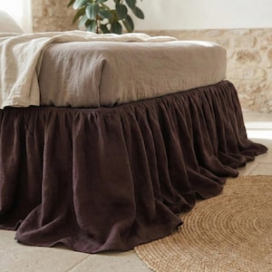 May include: A dark brown bed skirt with a gathered, ruffled design that hangs from the base of a bed. The bed is covered with a light beige fitted sheet and a matching light beige throw blanket draped over the top corner of the mattress.