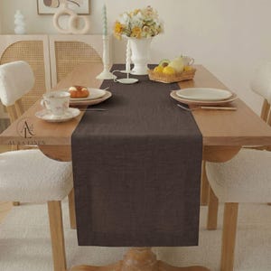 May include: A dark brown rectangular linen table runner centered on a light wood dining table. The runner features a simple hemmed edge. The table is set with white ceramic plates, a floral teacup, a wicker basket of fruit, and a white candle.
