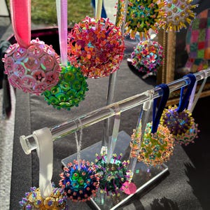 Peut inclure: Une collection d'ornements sphériques suspendus à un présentoir en acrylique transparent. Chaque ornement est recouvert de paillettes et de perles colorées, avec des motifs floraux dans des tons rose, vert, bleu et doré, suspendus par des rubans.