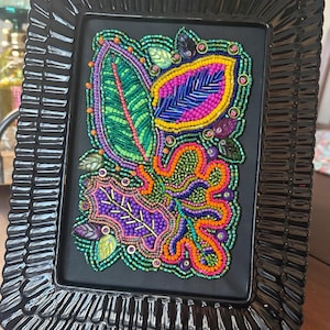 May include: A framed beaded artwork featuring colorful leaf designs. The leaves are adorned with beads in shades of green, purple, orange, and blue. The frame is black and has a textured, ridged design.