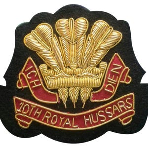 Könnte beinhalten: Ein schwarzer, gestickter Aufnäher mit goldenen Fäden. Das Design zeigt drei stilisierte goldene Federn über einem roten Banner mit dem Text "ICH DIEN" und einem unteren Banner mit der Aufschrift "10TH ROYAL HUSSARS".