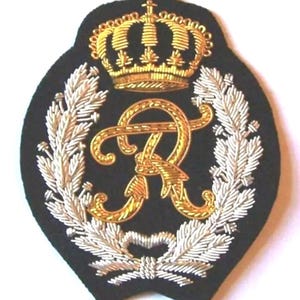 May include: Embroidered patch with a black background, featuring a gold crown, a gold monogram letter "R", and silver and gold laurel leaves. The patch has a unique, curved shape.