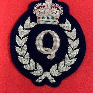 Pode incluir: Um patch bordado azul marinho com um "Q" prateado dentro de uma coroa de louros. Uma coroa prateada detalhada com detalhes vermelhos, verdes e azuis fica acima da coroa. O patch está sobre um fundo vermelho.