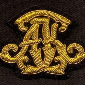 Op de afbeelding: Een goudkleurig, sierlijk monogram op een zwarte achtergrond. Het monogram bevat verweven letters met een getextureerd, touwachtig ontwerp. De algehele esthetiek is klassiek en elegant.