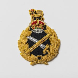 May include: A gold and black embroidered emblem featuring a crown, crossed swords, and a lion. The emblem is surrounded by a gold wreath and has intricate detailing. The background is white.