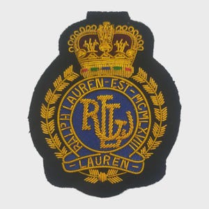 Puede incluir: Parche bordado con fondo negro, con una corona dorada y una corona de laurel. El centro tiene un círculo azul con letras doradas que dice "RL" y "Ralph Lauren".