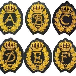 May include: Set of six embroidered patches, each featuring a gold crown atop a letter (A, B, C, D, E, F) within a gold laurel wreath on a black oval background. The patches are likely for decorative purposes.