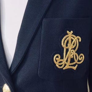 Puede incluir: Primer plano de una blazer azul marino con un emblema dorado bordado en el bolsillo. La blazer tiene un diseño clásico con solapa de muesca y un botón dorado. La tela parece ser un tejido texturizado.