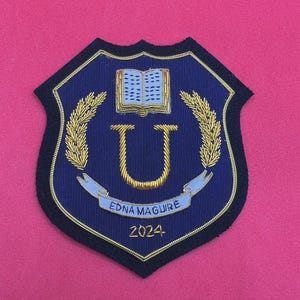 May include: A navy blue embroidered patch with a gold border and a shield shape. The patch features a gold letter "U", a banner with the name "Edna Maguire", and the year "2024". A gold open book and wheat stalks are also included.