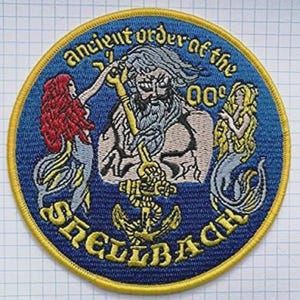 May include: Embroidered patch with a blue background and gold trim. The patch features the text "Ancient Order of the Shellbacks" and a central image of a bearded man holding a key, flanked by mermaids. An anchor is below the central figure.