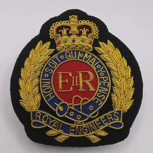 Op de afbeelding: Een zwart geborduurd embleem met een gouden kroon, laurierbladeren en tekst. Het centrale ontwerp omvat een rode cirkel met de letters "ER" en de woorden "HONI SOIT QUI MAL Y PENSE" en "ROYAL ENGINEERS" in blauw.