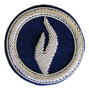 May include: A circular patch with a dark blue background and a silver flame design in the center. The flame is surrounded by a silver, braided border. The outer edge of the patch has a silver rope-like trim.