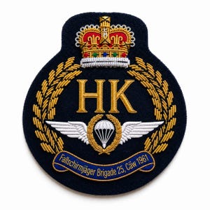 May include: A navy blue embroidered patch featuring a gold crown, the letters "HK", and a parachute with wings. The patch includes the text "Fallschirmjäger Brigade 25, Cäw 1961" on a blue banner, surrounded by gold laurel leaves.