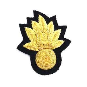 May include: A gold embroidered patch with a black felt backing. The patch features a circular base with a radiating design, topped by a fan-shaped arrangement of gold embroidery. The black felt backing is cut in a starburst shape.