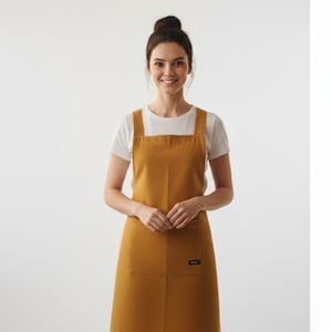 Può includere: Grembiule color senape con un'ampia tasca frontale e una piccola etichetta nera. Il grembiule ha un design semplice e minimalista con scollatura quadrata e ampie spalline. La modella indossa una maglietta bianca sotto.