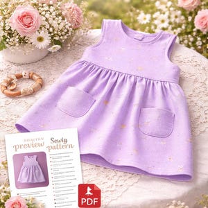 May include: A lavender baby dress with two front pockets and gold accents. The dress is displayed on a table with a lace tablecloth, surrounded by flowers and a sewing pattern preview. The pattern is for 0-3 months.