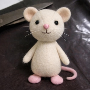 May include: A handmade, off-white felt mouse with pink accents. The plush toy features black button eyes, a pink nose, and whiskers. The mouse has pink feet and a curled pink tail. A charming, whimsical stuffed animal.