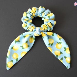 Pode incluir: Uma scrunchie azul claro com um padrão de limão amarelo e um laço a condizer. O acessório de cabelo é feito de tecido e foi concebido para segurar o cabelo. O laço adiciona um elemento decorativo.