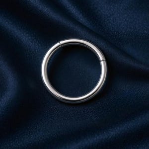 Könnte beinhalten: Ein silberfarbener, kreisförmiger Klappring, wahrscheinlich ein Schmuckstück, wird vor einem dunkelblauen, strukturierten Hintergrund gezeigt. Die Oberfläche des Rings ist glatt und reflektierend, mit einem sichtbaren Scharnier. Die Gesamtästhetik ist minimalistisch und modern.