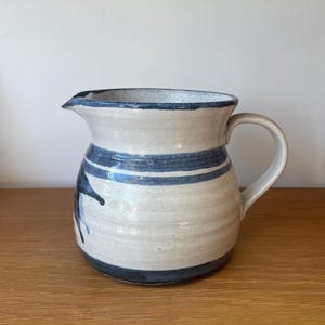 May include: A ceramic jug with a cream-coloured body and blue accents. The jug features a dark blue rim and two horizontal stripes. A blue abstract design is painted on the side. The jug has a handle and a spout.