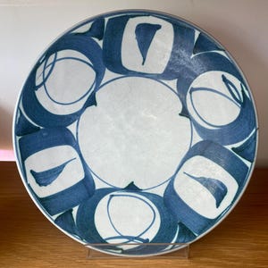 May include: A white ceramic plate with a blue abstract design. The plate features a central white circle surrounded by blue shapes and patterns. The plate is displayed on a clear stand.