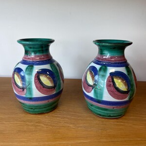 May include: Two ceramic vases with a rounded body and a flared rim. The vases feature a hand-painted pattern of green vertical stripes, horizontal blue and pink bands, and circular motifs in red, yellow, and blue against a white background.