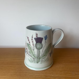 May include: A ceramic mug decorated with a thistle motif. The mug has a light blue interior and a handle. The thistle is illustrated with purple, green, and blue colours. The mug is on a wooden surface.