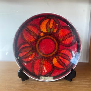 May include: A decorative plate with a floral design. The plate has a red base with orange and black accents. The floral pattern is centered, with a clear rim. The plate is displayed on a black stand. The plate's diameter is approximately 20 cm.