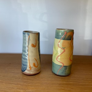 May include: Two cylindrical ceramic vases with hand-painted designs. The left vase features blue and cream tones with orange accents. The right vase displays green and yellow horizontal bands with a wavy orange line running vertically across the surface.
