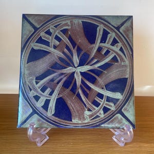 May include: Square ceramic tile with a blue background and an abstract design in shades of brown and teal. The design features a central floral-like pattern surrounded by curved lines. The tile is displayed on a clear plastic stand.