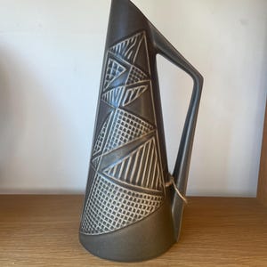 May include: A dark brown ceramic jug with a geometric pattern. The jug has a tall, angled body with a handle and is decorated with textured triangles, lines, and dots. A small tag is attached to the handle.