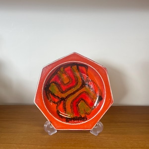 May include: An octagonal orange ceramic dish with a central abstract design in red, brown, and green. The dish is displayed on a clear plastic stand. The dish's design and shape suggest it is a vintage piece, adding to its appeal.