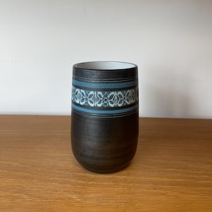 May include: A cylindrical ceramic tumbler with a matte black exterior finish. The upper section features a decorative band with horizontal blue stripes and a repeating white geometric pattern. The interior of the vessel is white.