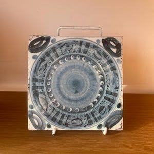 May include: Square ceramic trivet with a blue and white circular design. The trivet has a metal handle and metal feet. The design features a central circle with dotted and leaf-like patterns. The trivet is on a wooden surface.
