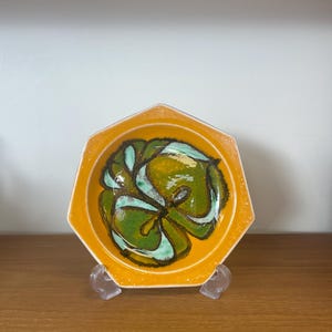 May include: A hexagonal ceramic dish with a vibrant yellow exterior and a central design of green and teal abstract shapes. The dish is displayed on a clear plastic stand, highlighting its unique design and colour palette.