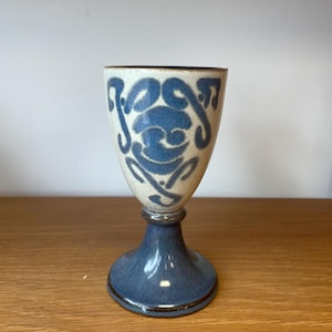 May include: A ceramic goblet featuring a cream-coloured bowl with a blue hand-painted abstract pattern and a solid blue glazed stem and base. The vessel has a smooth finish and a flared rim, standing on a circular pedestal foot with a dark rim.