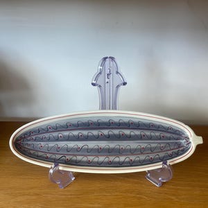 May include: An oval ceramic serving dish with a white rim and a grey interior. The interior features a pattern of wavy black lines and red dots. The dish is displayed on a clear plastic stand.