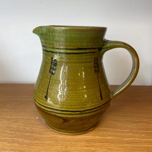 May include: A ceramic pitcher with a glossy olive green glaze, featuring a rounded body, a curved handle, and a pouring spout. The exterior is decorated with horizontal dark brown bands and vertical sprigs of wheat motifs in dark brown paint.