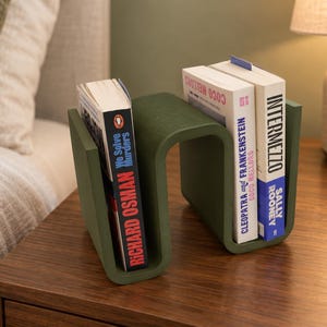 Peut inclure: Un porte-livres en forme de S vert olive mat contenant trois livres. Le côté gauche contient We Solve Murders de Richard Osman. Le côté droit contient Cleopatra and Frankenstein de Coco Mellors et Intermezzo de Sally Rooney, tous debout.