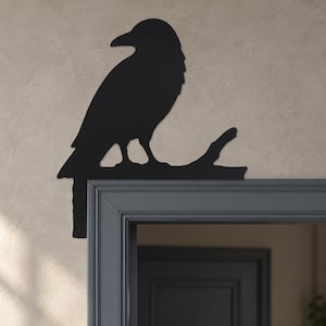 Pode incluir: Uma decoração de parede em metal preto fosco em forma de corvo pousado num galho, projetada para ser montada no canto superior da moldura de uma porta. A silhueta mostra o pássaro virado para a esquerda com as penas da cauda para baixo.