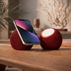 Könnte beinhalten: Ein rotes, kugelförmiges kabelloses Ladegerät und Lautsprecherset. Das Ladegerät hält ein Smartphone mit farbenfrohem Display. Der Lautsprecher hat eine weiße Oberseite. Beide Geräte haben eine gerippte Textur und stehen auf einer Holzoberfläche.
