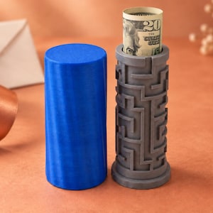 Könnte beinhalten: Ein zylindrischer blauer Behälter neben einer grauen Puzzle-Box mit einem Labyrinth-Muster auf der Außenseite. Die graue Puzzle-Box ist oben offen und zeigt einen aufgerollten Zwanzig-Dollar-Schein, der im hohlen Zentrum steckt.