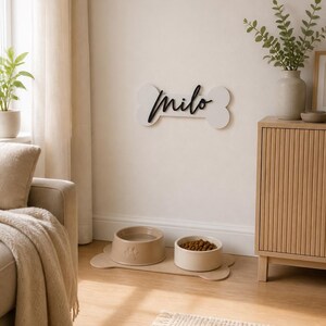 May include: A white wall-mounted bone-shaped sign with the black script name Milo, paired with a beige bone-shaped floor mat holding two round bowls, one containing water and the other filled with brown dry pet food kibble.