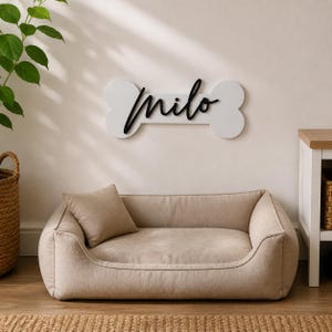 Può includere: Una cuccia rettangolare per animali color beige con un cuscino quadrato abbinato, posizionata sotto una targa da parete bianca a forma di osso di cane. La targa presenta il nome "milo" in lettere corsive nere e tridimensionali.