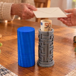 Könnte beinhalten: Ein zylindrischer blauer Behälter neben einer grauen zylindrischen Puzzle-Box. Die graue Puzzle-Box weist ein komplexes Labyrinth-Muster auf der Außenseite auf und enthält einen gerollten 50-Euro-Schein, der an der oberen Öffnung sichtbar ist.