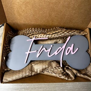 Personalisiertes Hunde-Namensschild Frida auf grauem Knochen mit rosa 3D-Schriftzug, verpackt in einer Geschenkbox.