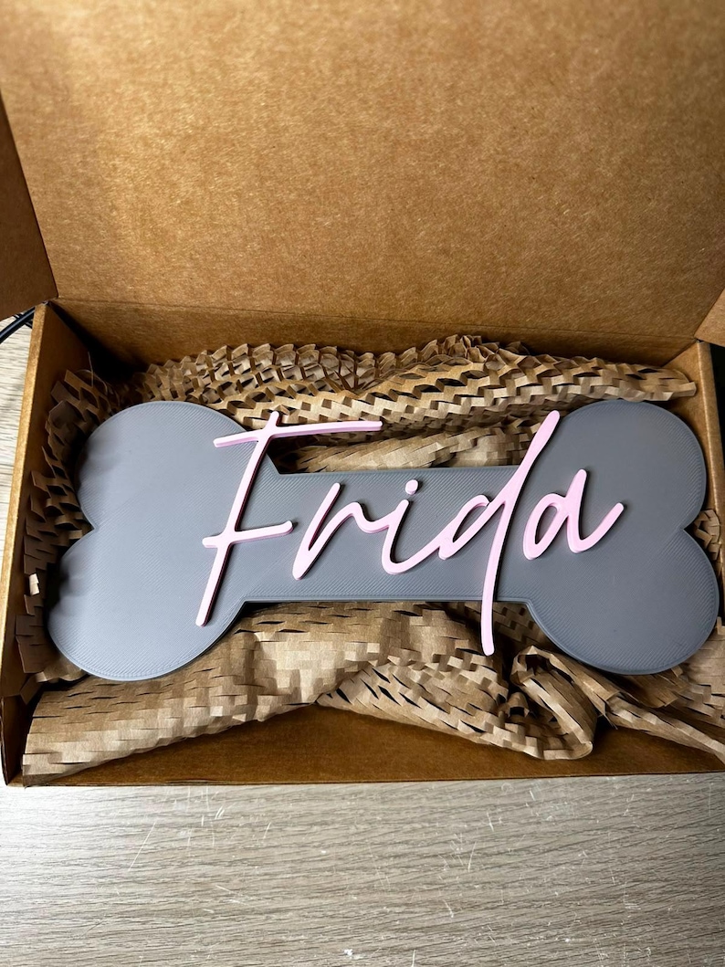 Personalisiertes Hunde-Namensschild Frida auf grauem Knochen mit rosa 3D-Schriftzug, verpackt in einer Geschenkbox.
