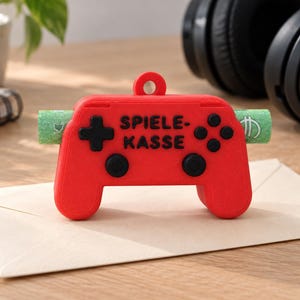 Könnte beinhalten: Eine leuchtend rote Spardose in Form eines Game-Controllers mit einer kleinen runden Öse an der Oberseite. Die Vorderseite zeigt schwarze Tasten und den Schriftzug "SPIELE-KASSE" in schwarzen Buchstaben. Ein grüner Geldschein steckt horizontal darin.